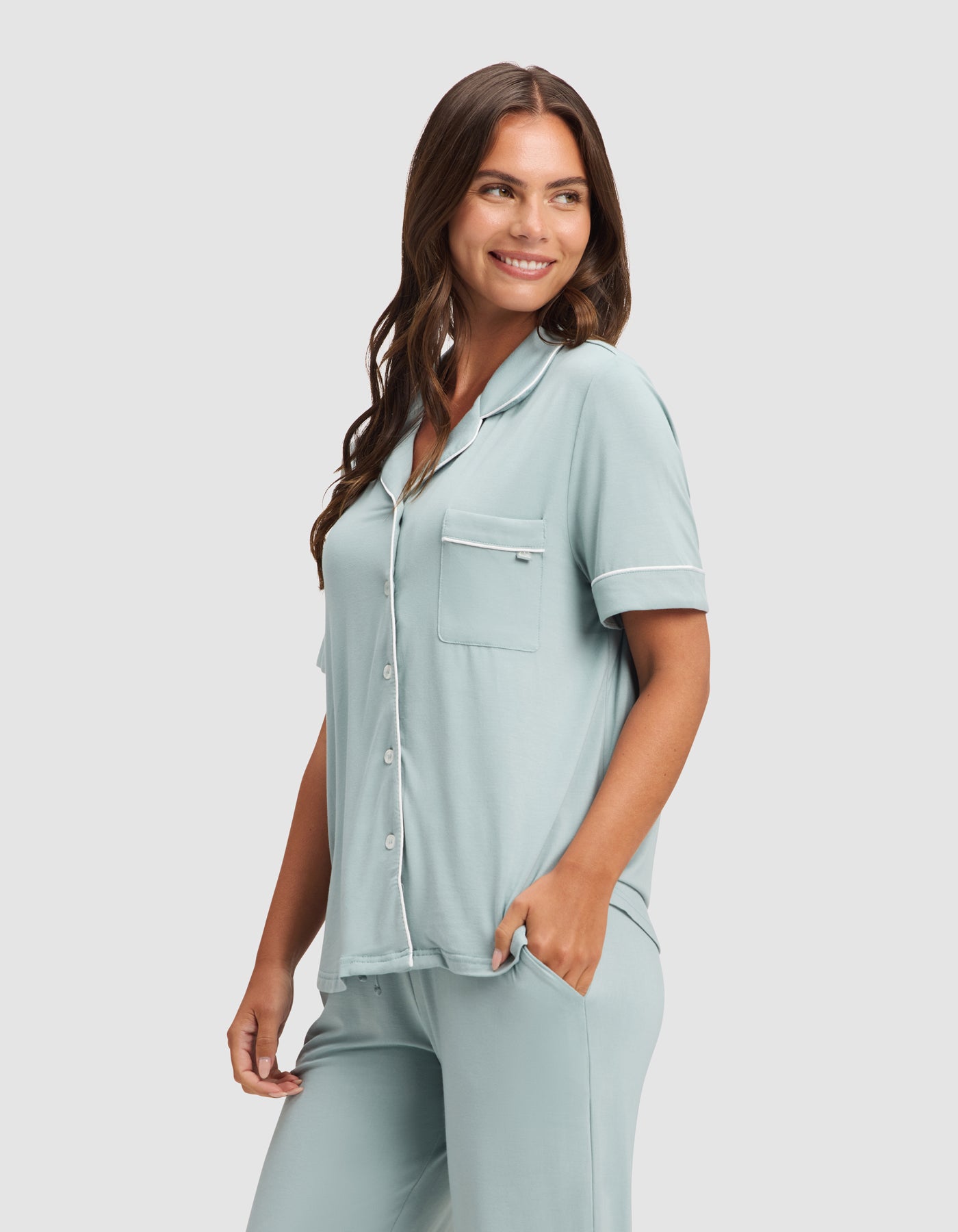 A woman smiles with one hand in her pocket, wearing the Cozy Earth Women's Bamboo Stretch-Knit Short Sleeve & Pant Pajama Set in light blue, featuring a button-up top and matching pants against a plain light background. 