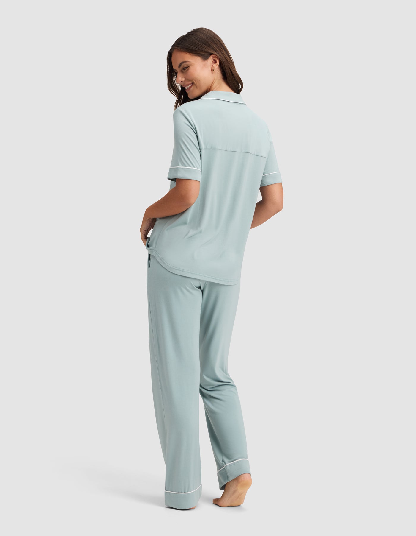 A woman, barefoot and smiling over her shoulder, models the Cozy Earth Women's Bamboo Stretch-Knit Short Sleeve & Pant Pajama Set in light blue with white trim against a simple background. 