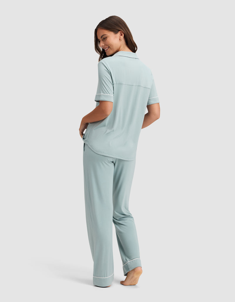 A woman, barefoot and smiling over her shoulder, models the Cozy Earth Women's Bamboo Stretch-Knit Short Sleeve & Pant Pajama Set in light blue with white trim against a simple background.