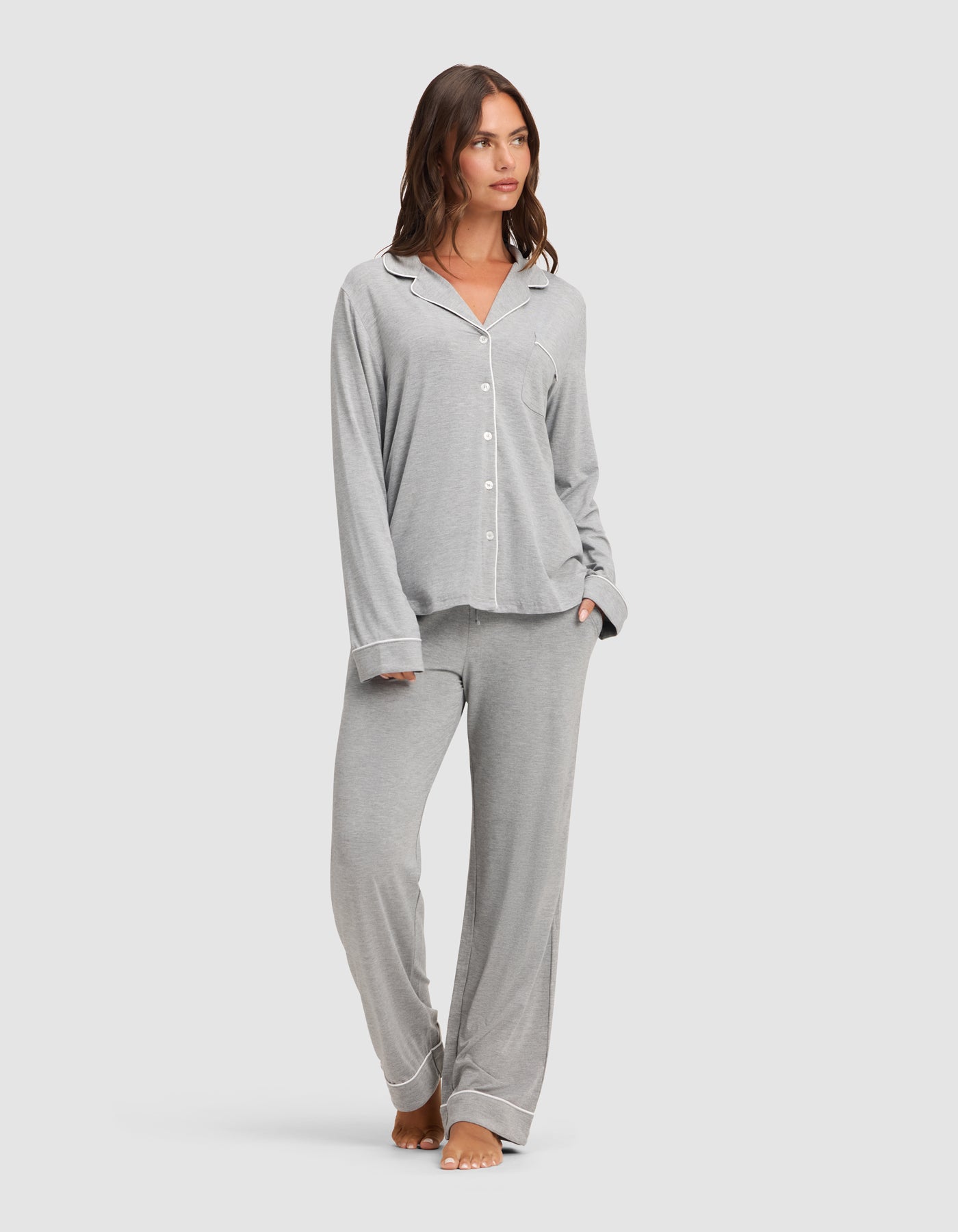 A woman stands barefoot against a plain light background, wearing the Cozy Earth Women's Bamboo Stretch-Knit Long Sleeve Pajama Set in light grey with white piping, featuring a button-up top and matching pants.  