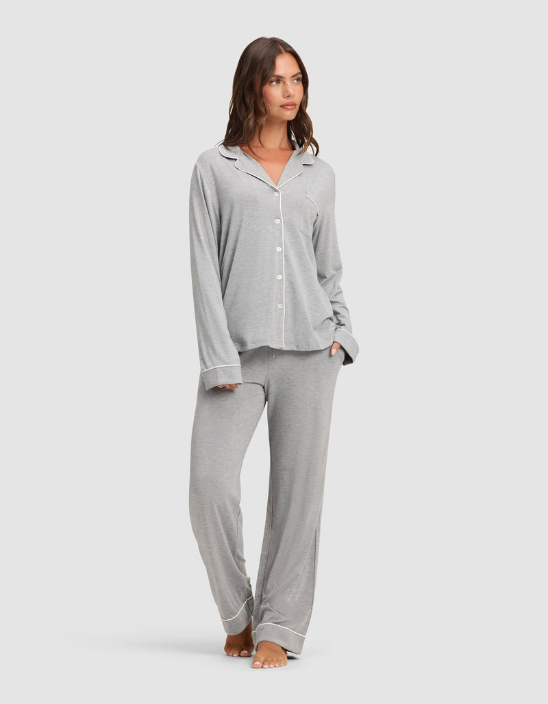 A woman stands barefoot against a plain light background, wearing the Cozy Earth Women's Bamboo Stretch-Knit Long Sleeve Pajama Set in light grey with white piping, featuring a button-up top and matching pants.