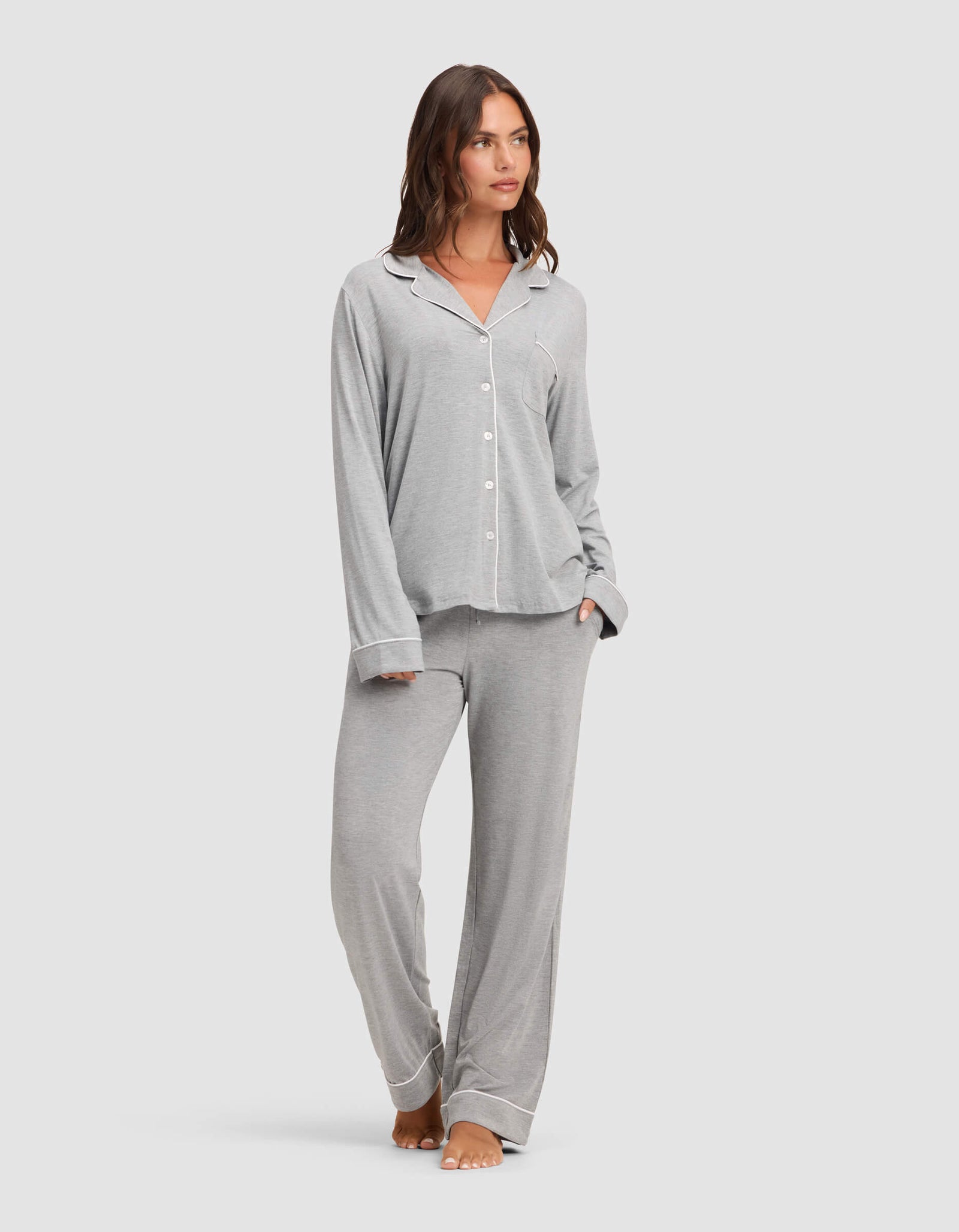 A woman stands barefoot against a plain light background, wearing the Cozy Earth Women's Bamboo Stretch-Knit Long Sleeve Pajama Set in light grey with white piping, featuring a button-up top and matching pants.  