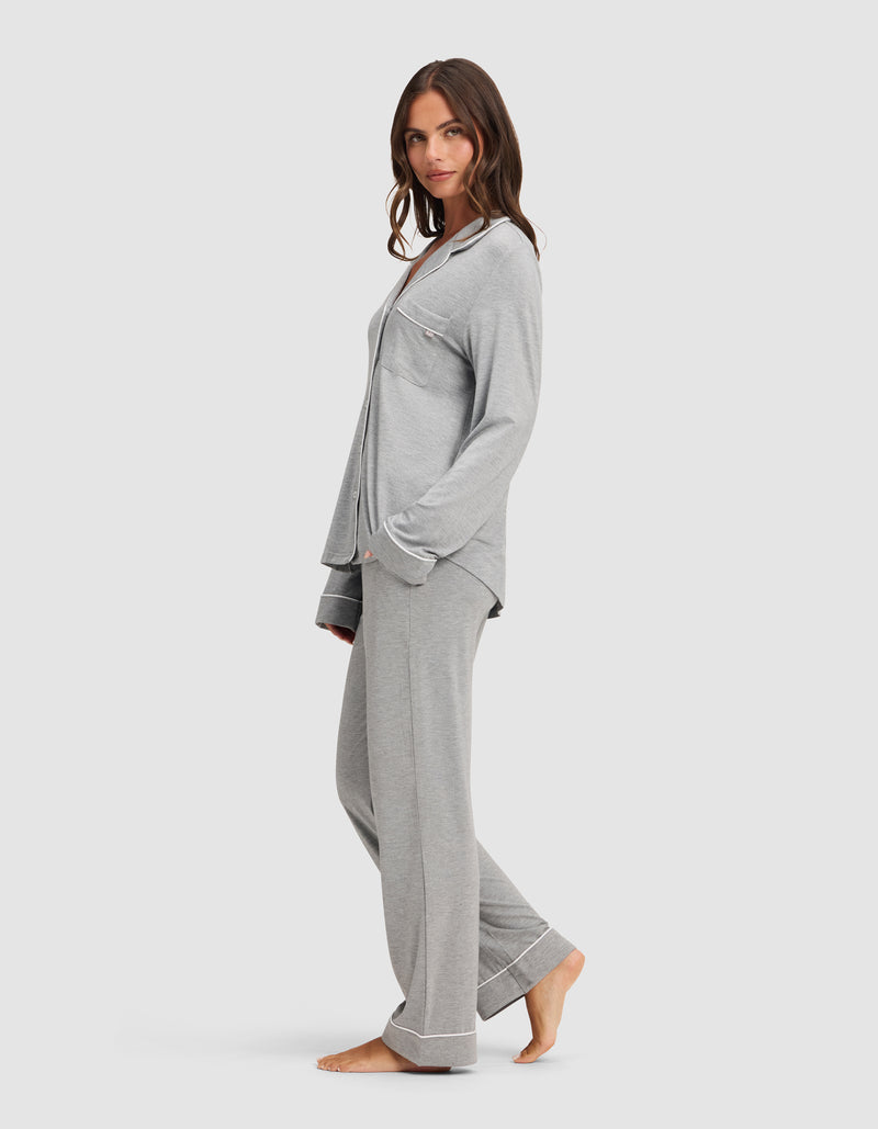 A woman stands barefoot in a relaxed pose, wearing the Cozy Earth Women's Bamboo Stretch-Knit Long Sleeve Pajama Set in light gray with button-up top and chest pocket, against a plain white background.