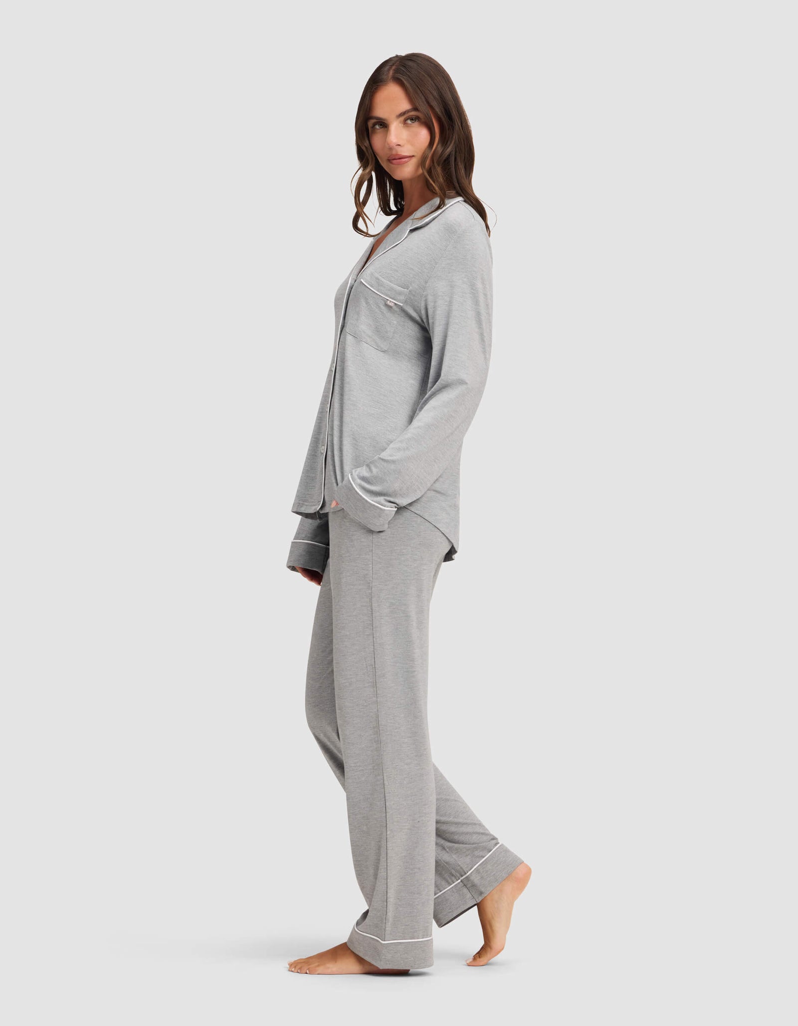 A woman stands barefoot in a relaxed pose, wearing the Cozy Earth Women's Bamboo Stretch-Knit Long Sleeve Pajama Set in light gray with button-up top and chest pocket, against a plain white background. 