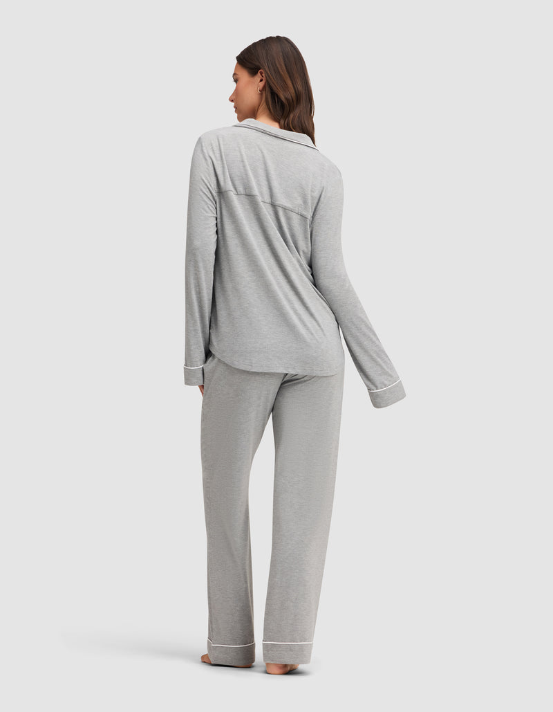 A woman stands barefoot, facing away, wearing the Cozy Earth Women's Bamboo Stretch-Knit Long Sleeve Pajama Set in light gray with white trim. She is on a plain light background.