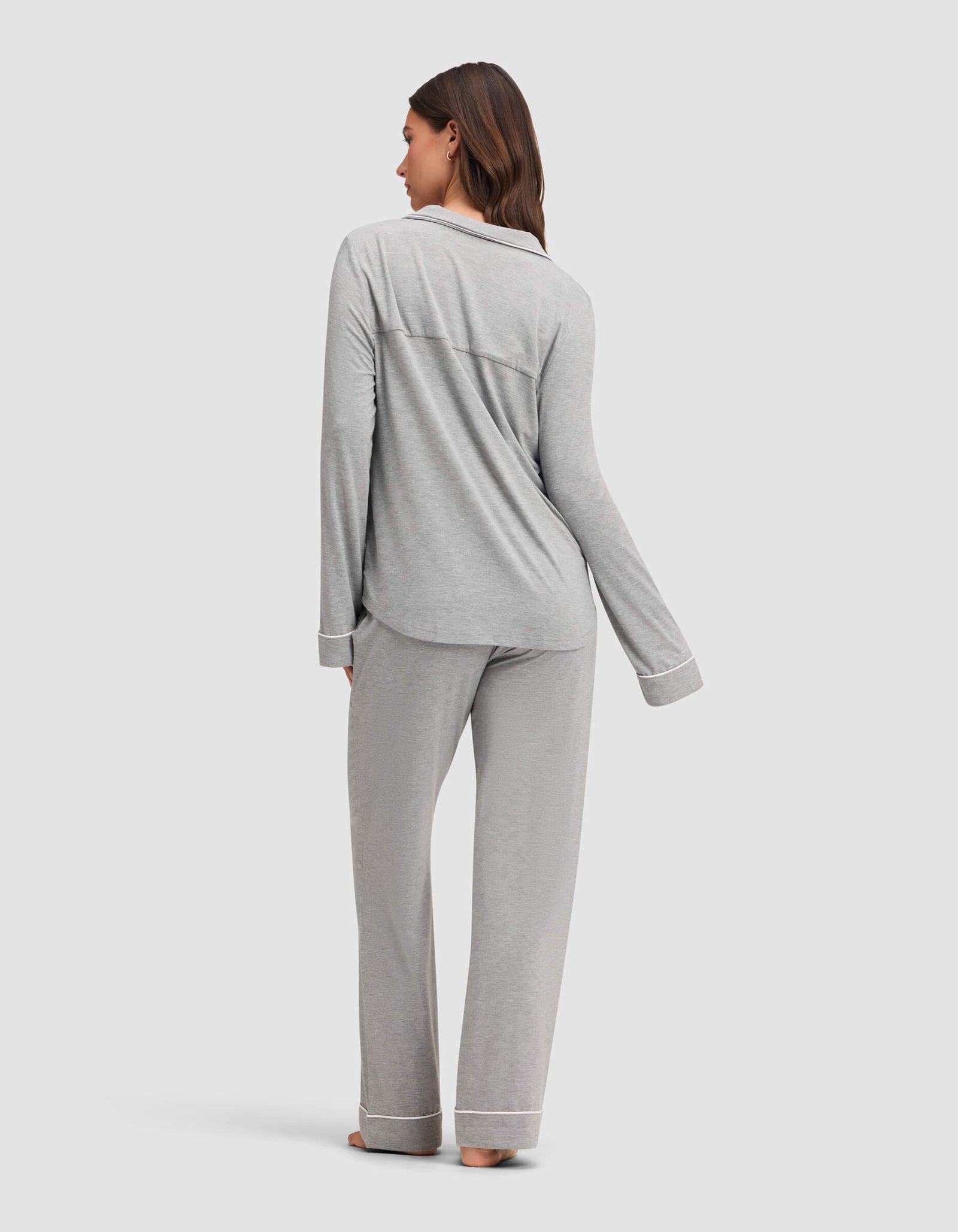 A woman stands barefoot, facing away, wearing the Cozy Earth Women's Bamboo Stretch-Knit Long Sleeve Pajama Set in light gray with white trim. She is on a plain light background. 