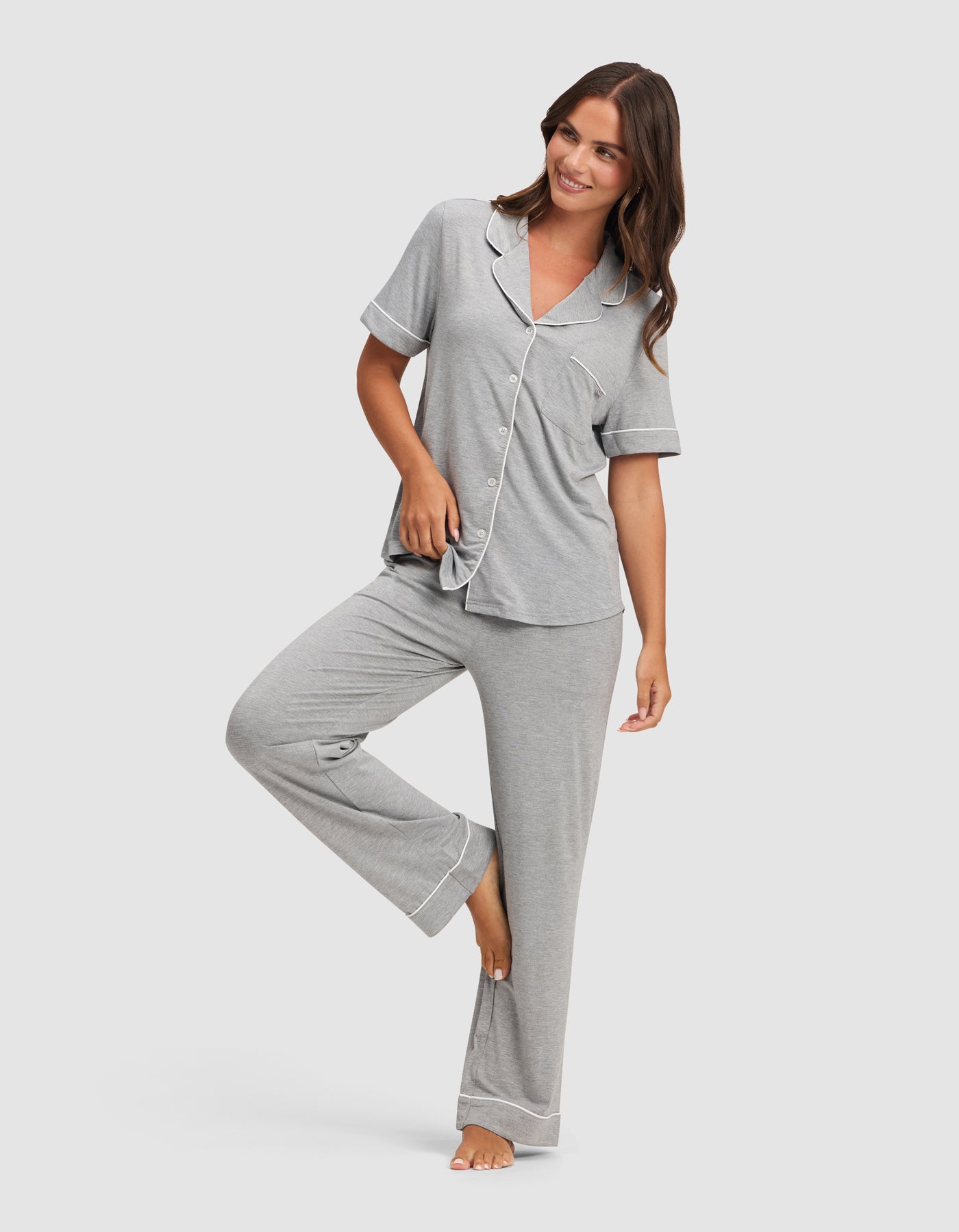 A woman models the Cozy Earth Women's Bamboo Stretch-Knit Short Sleeve & Pant Pajama Set in light gray with white trim, standing barefoot, smiling, and lifting one leg as she holds the hem of her top. 