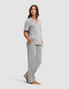 A woman stands barefoot in a Cozy Earth Women's Bamboo Stretch-Knit Short Sleeve & Pant Pajama Set, featuring light gray fabric with white piping and loose pants, hands in pockets, against a plain light background.  |Color:Grey