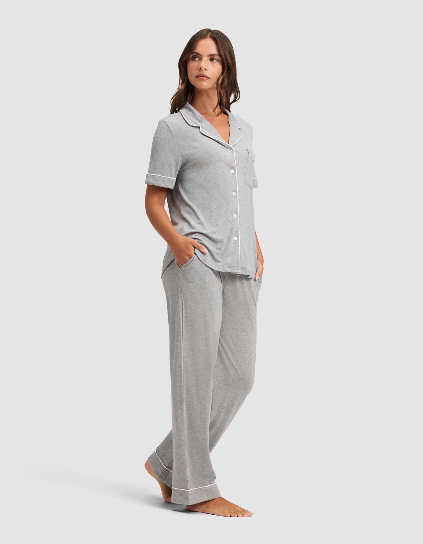 A woman stands barefoot in a Cozy Earth Women's Bamboo Stretch-Knit Short Sleeve & Pant Pajama Set, featuring light gray fabric with white piping and loose pants, hands in pockets, against a plain light background.  