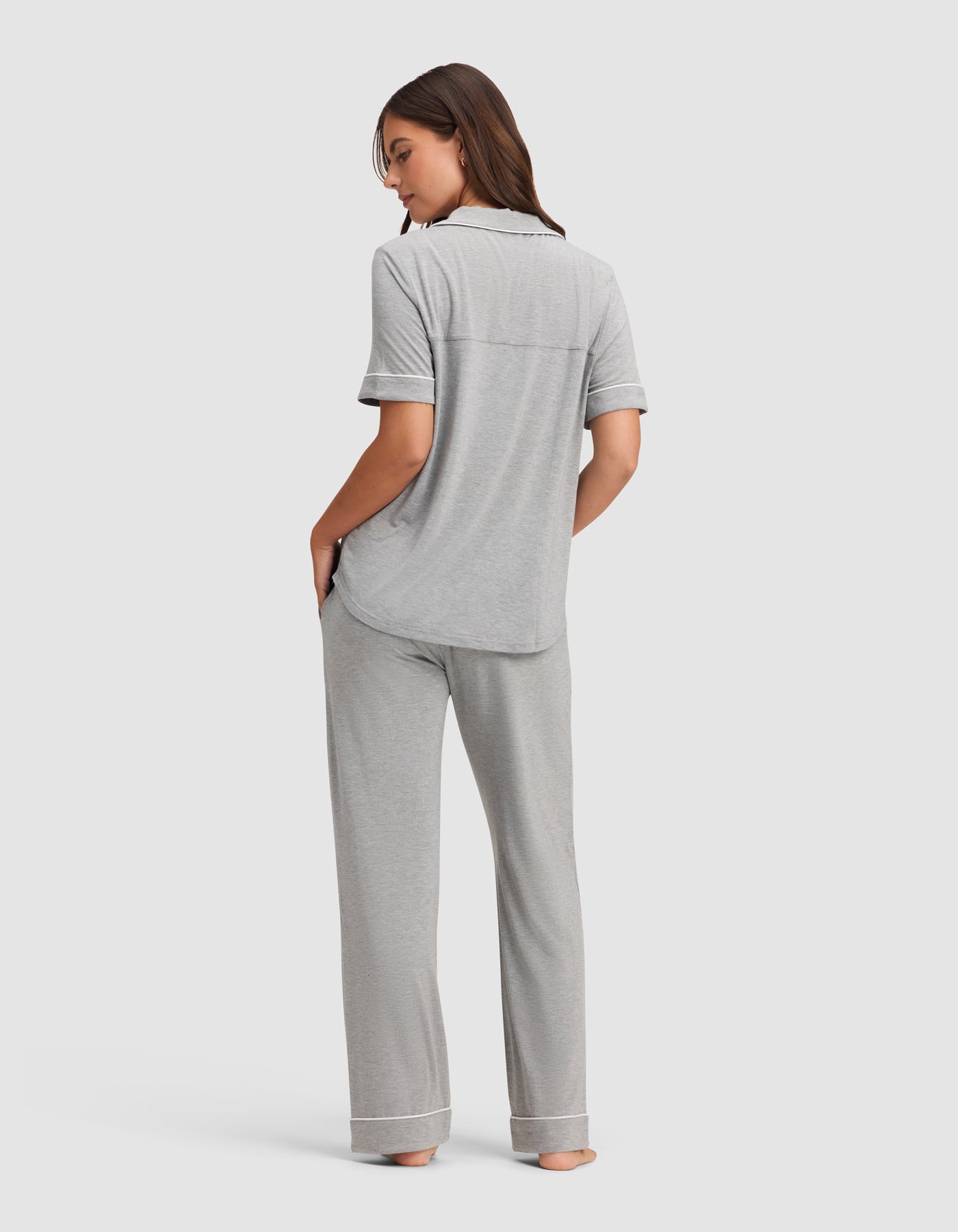 A woman stands barefoot, facing away, wearing the Cozy Earth Women's Bamboo Stretch-Knit Short Sleeve & Pant Pajama Set in light gray, featuring cuffed long pants. She has one hand in her pocket and her head turned slightly to the side. 