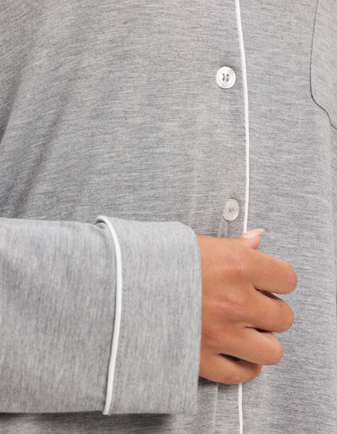 A close-up of a person’s hand holding the front of Cozy Earth’s Women’s Bamboo Stretch-Knit Long Sleeve Pajama Set in light grey, featuring white piping and buttons. The soft bamboo fabric looks comfortable and cozy. 