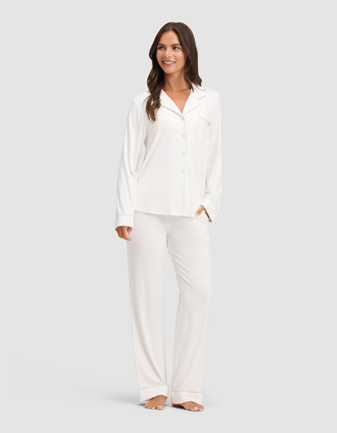 A woman smiles while wearing the Cozy Earth Women's Bamboo Stretch-Knit Long Sleeve Pajama Set, featuring a button-up top. She stands barefoot against a plain light gray background. 