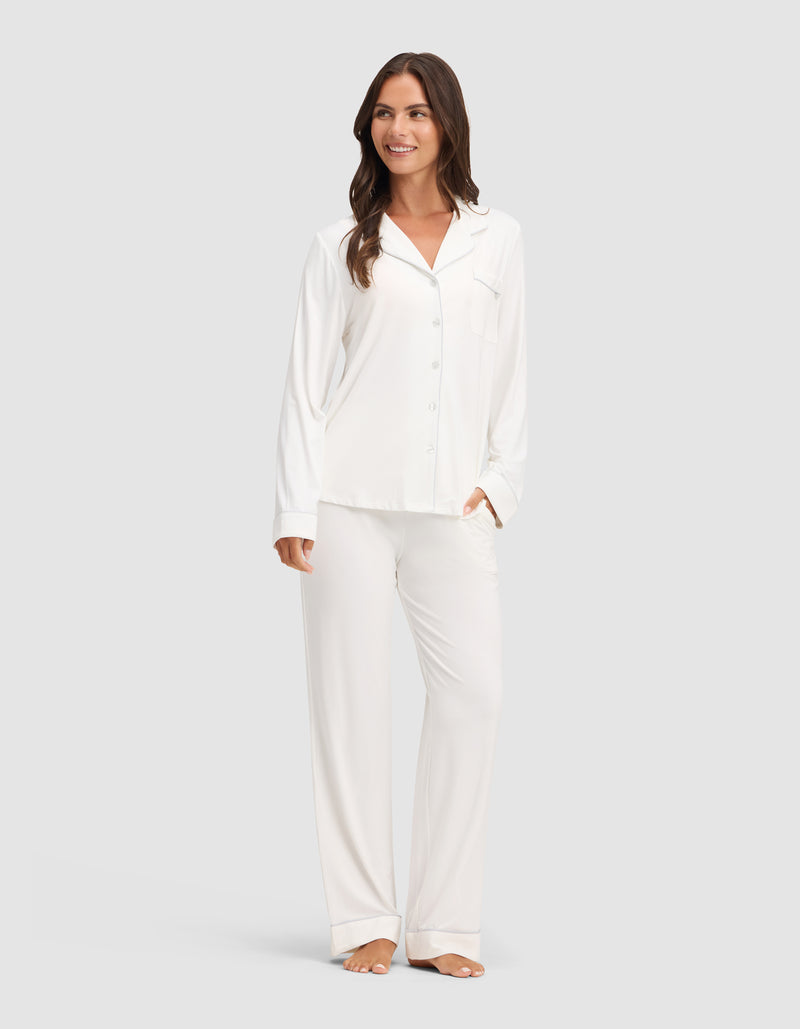 A woman smiles while wearing the Cozy Earth Women's Bamboo Stretch-Knit Long Sleeve Pajama Set, featuring a button-up top. She stands barefoot against a plain light gray background.