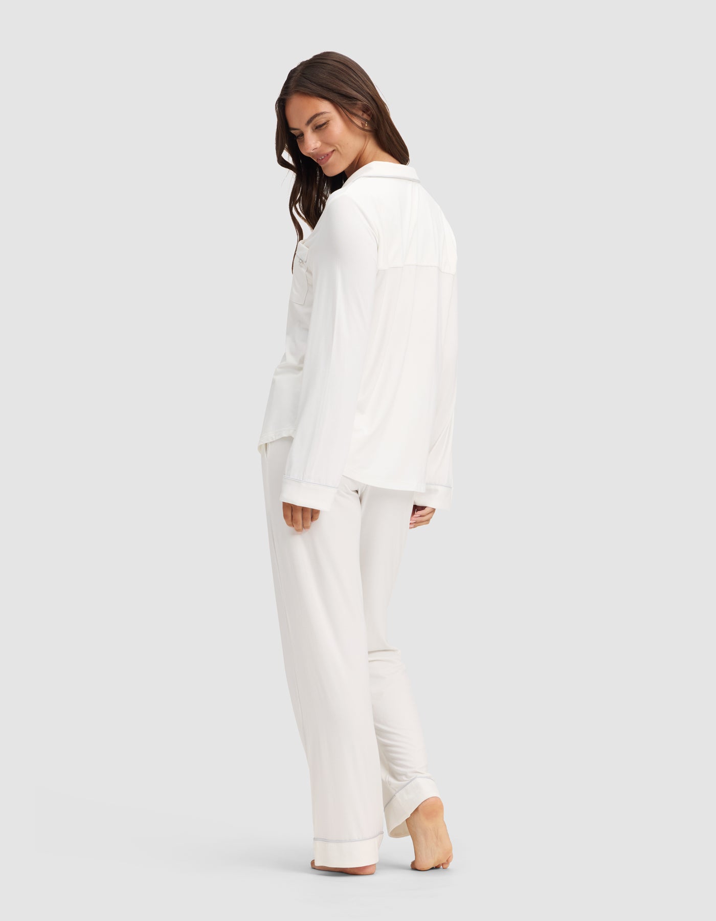 A woman with long brown hair stands barefoot, smiling and turning slightly to the side. She wears the Cozy Earth Women's Bamboo Stretch-Knit Long Sleeve Pajama Set in white, with pants, against a plain light gray background. 