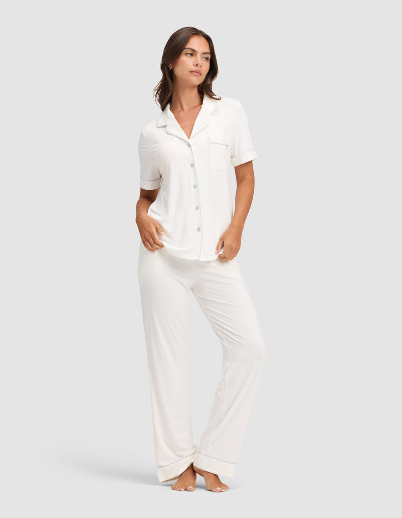A woman wears Cozy Earth's Women's Bamboo Stretch-Knit Short Sleeve & Pant Pajama Set, featuring a white short-sleeved button-up top with chest pocket and matching long pants, standing barefoot against a plain light gray background.