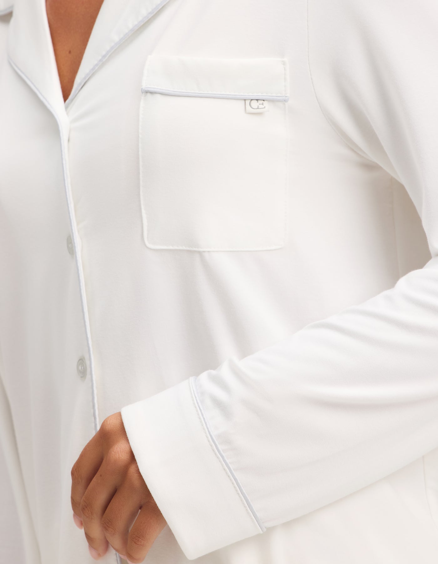 A close-up shows a person wearing the Cozy Earth Women's Bamboo Stretch-Knit Long Sleeve Pajama Set, featuring a white button-up top with a chest pocket and light piping. Only part of the torso and bent arm are visible. 