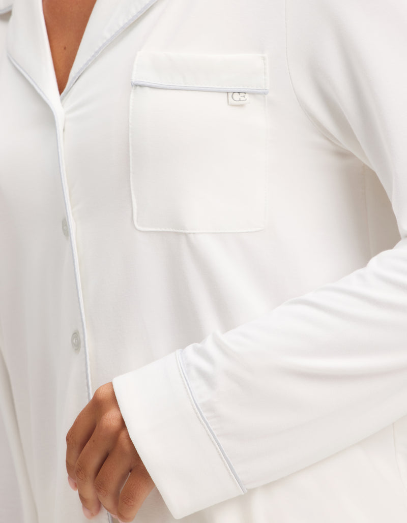 A close-up shows a person wearing the Cozy Earth Women's Bamboo Stretch-Knit Long Sleeve Pajama Set, featuring a white button-up top with a chest pocket and light piping. Only part of the torso and bent arm are visible.