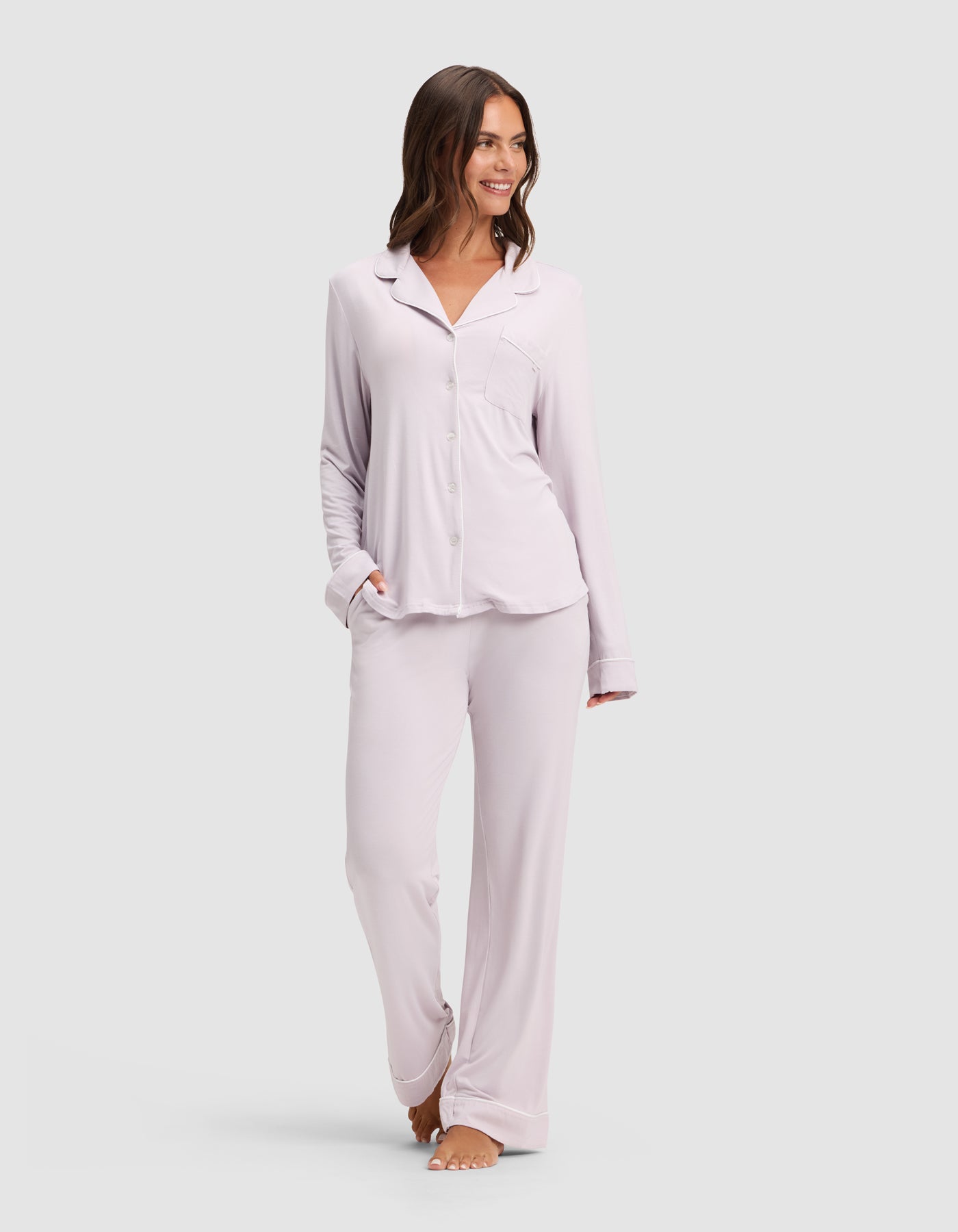 A woman smiles while wearing the Cozy Earth Women's Bamboo Stretch-Knit Long Sleeve Pajama Set, featuring a button-up top and matching pants in light lavender, standing barefoot against a plain light background. 