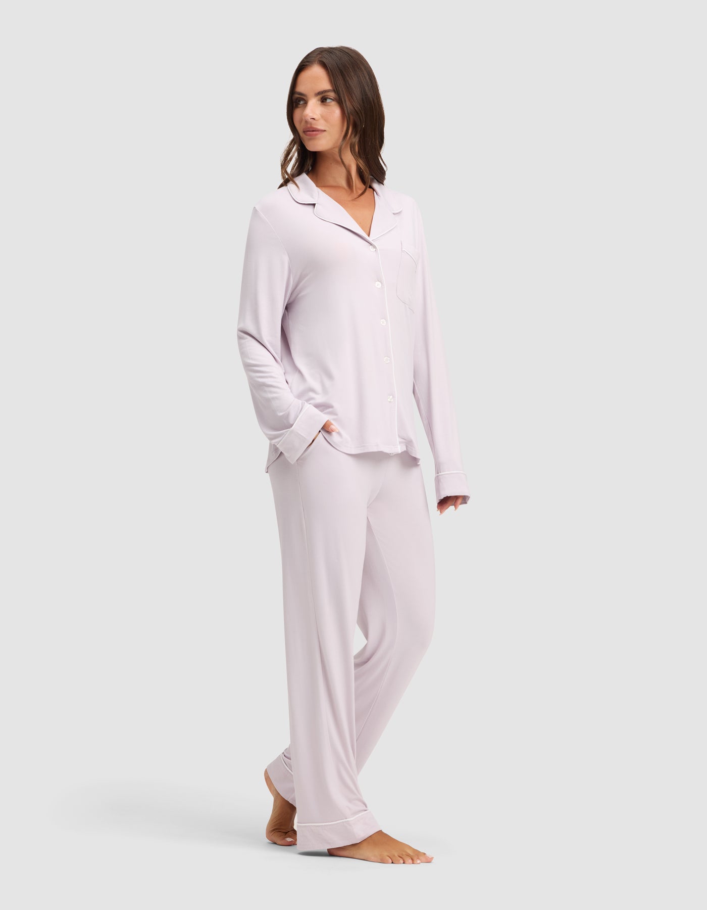 A woman wears the Cozy Earth Women's Bamboo Stretch-Knit Long Sleeve Pajama Set in light purple with matching pants, standing barefoot on a light gray background and looking to the side with one hand in her pocket. 
