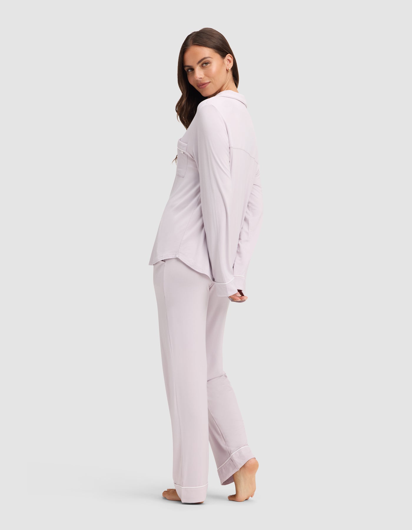 A woman stands barefoot on a light background, turned slightly to the side and looking over her shoulder. She wears the Cozy Earth Women's Bamboo Stretch-Knit Long Sleeve Pajama Set in matching light lavender. 