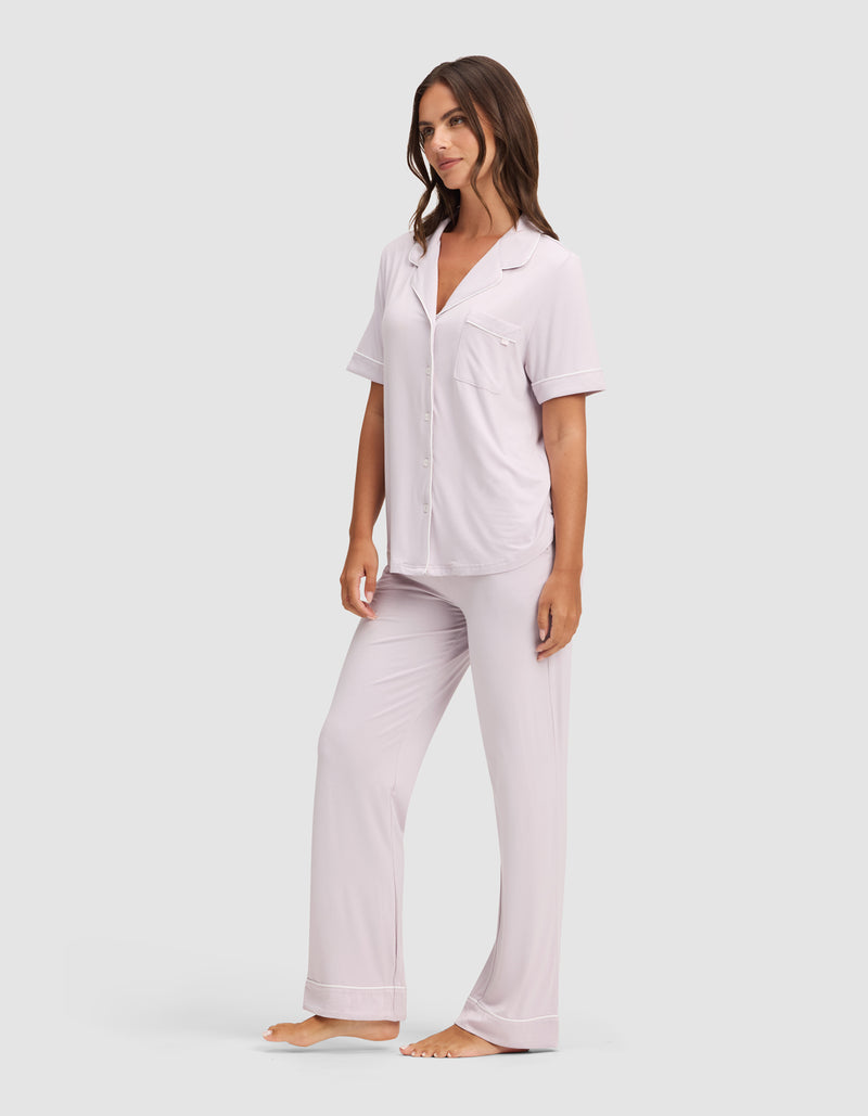 A woman stands barefoot in Cozy Earth's Women's Bamboo Stretch-Knit Short Sleeve & Pant Pajama Set, featuring a light pink button-up shirt and long pants, against a plain light gray background.