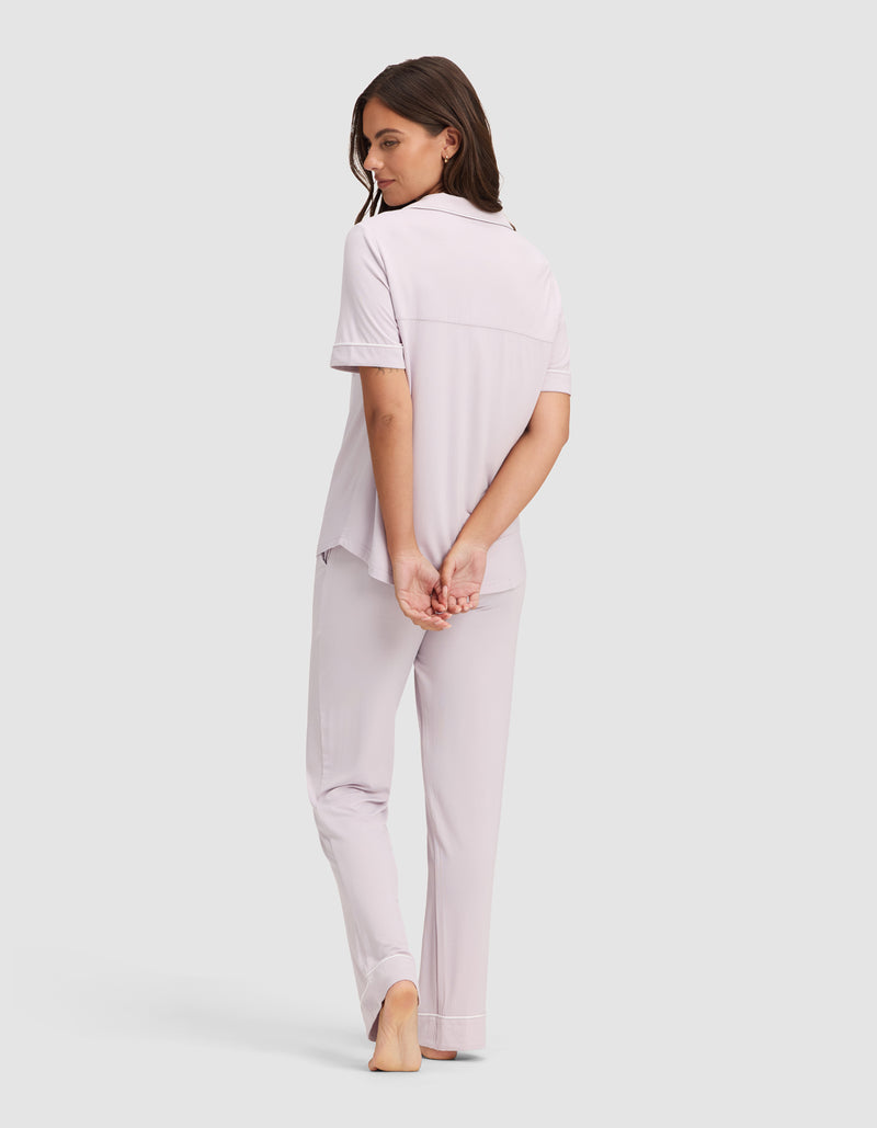 A woman models the Cozy Earth Women's Bamboo Stretch-Knit Short Sleeve & Pant Pajama Set in light lavender, standing barefoot with her hands behind her back, looking over her shoulder against a plain light background.