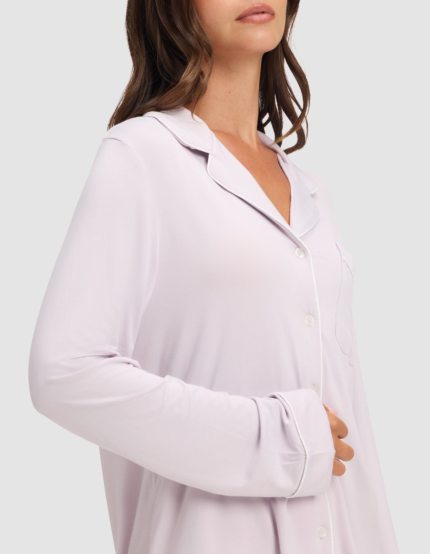 A woman wears the Cozy Earth Women's Bamboo Stretch-Knit Long Sleeve Pajama Set in light pink with white piping, standing against a plain background with one hand on her abdomen. Her face is partially out of frame. 