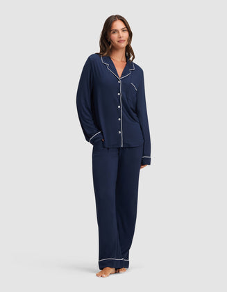 A woman poses barefoot against a plain light background, smiling in the Cozy Earth Women's Bamboo Stretch-Knit Long Sleeve Pajama Set—navy blue with white piping, featuring a button-up top and loose-fitting pants. |Color:Navy