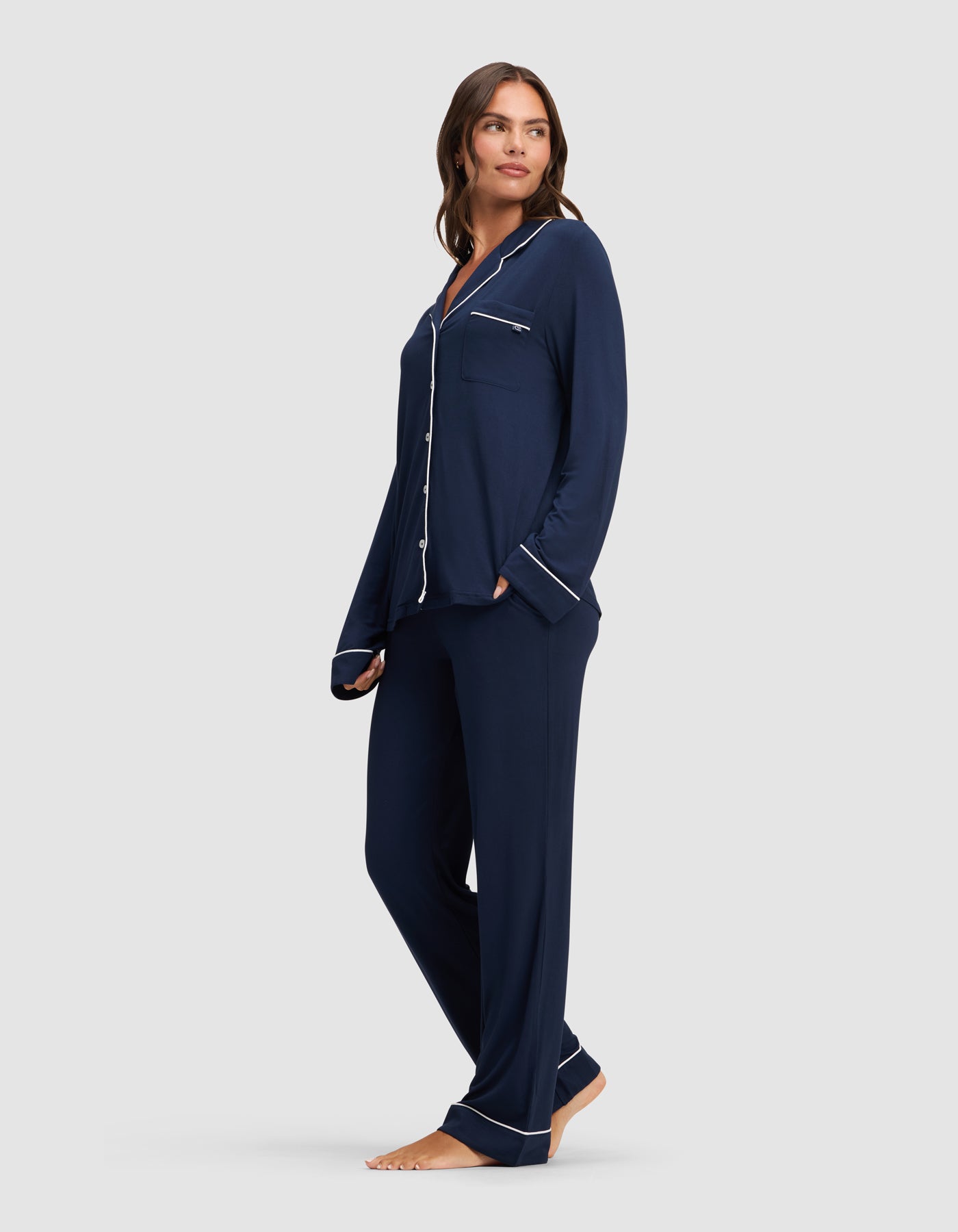 A woman stands barefoot against a plain light gray background, wearing the Cozy Earth Women's Bamboo Stretch-Knit Long Sleeve Pajama Set in navy with white piping, featuring a button-up top and matching pants.