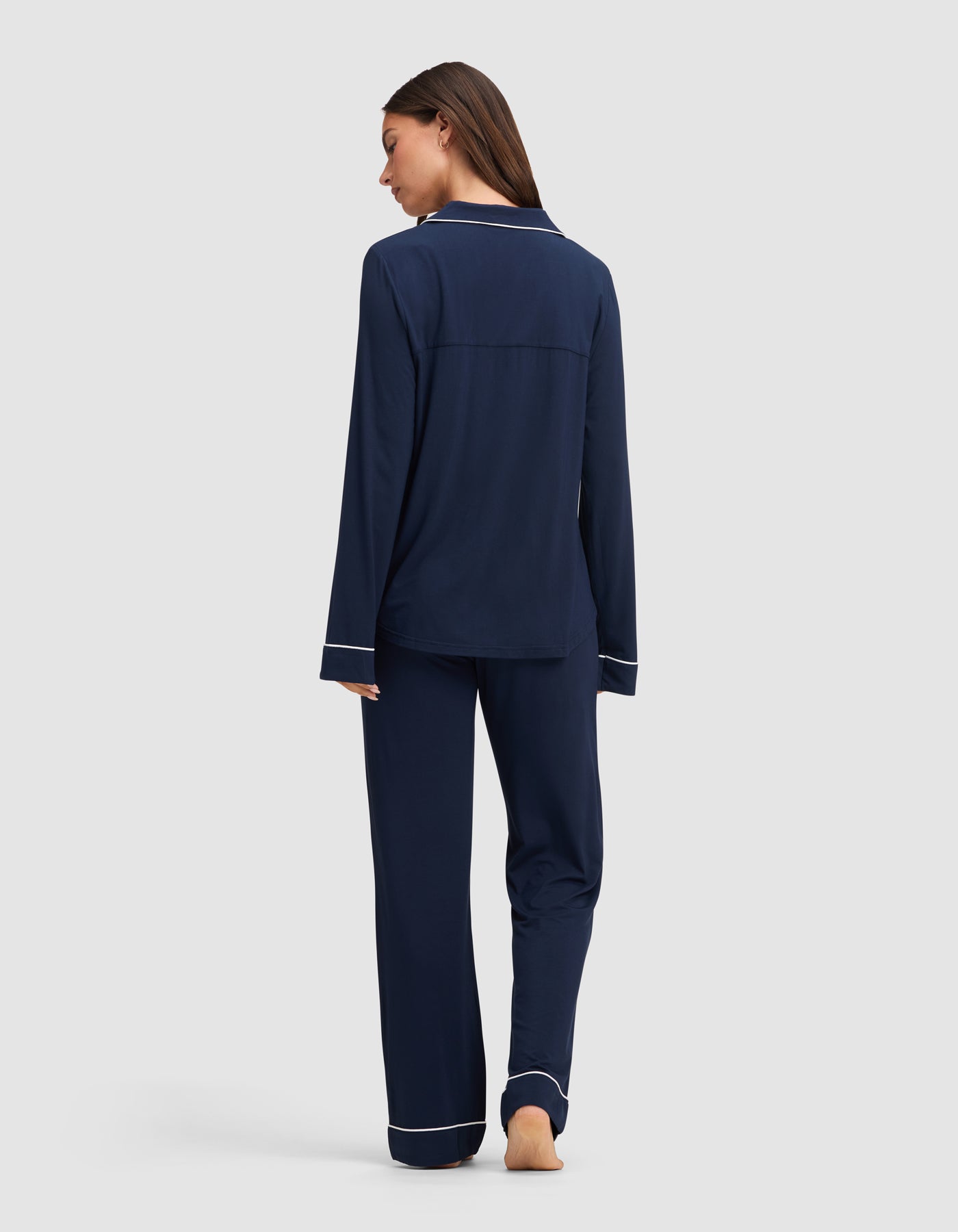 A woman, barefoot and facing away, wears the Cozy Earth Women's Bamboo Stretch-Knit Long Sleeve Pajama Set in navy blue with white piping on the cuffs and hem, set against a plain light gray background.