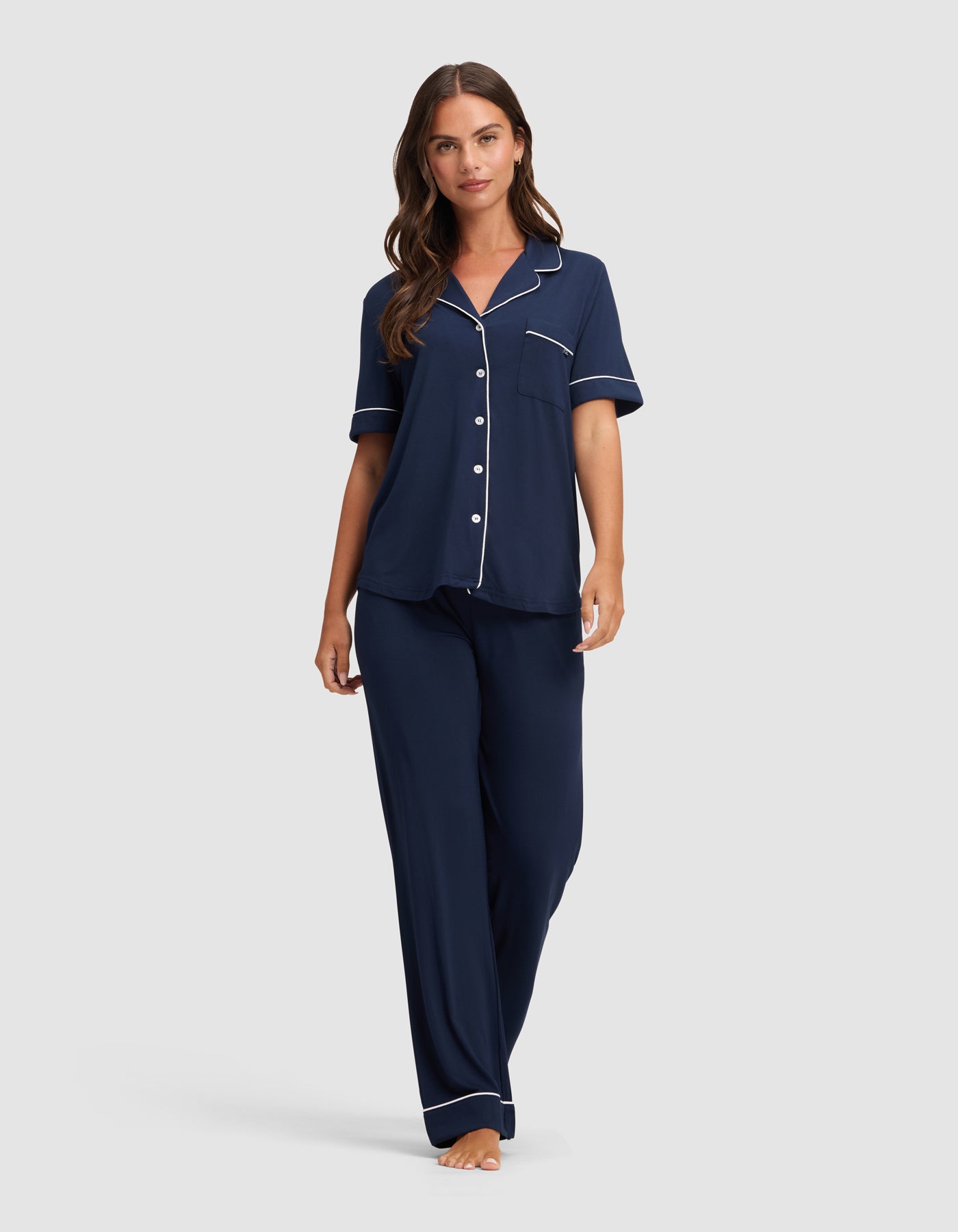 A woman wears the Cozy Earth Women's Bamboo Stretch-Knit Short Sleeve & Pant Pajama Set in navy blue with white piping, featuring a button-up top and long pants, while standing barefoot against a plain light background.