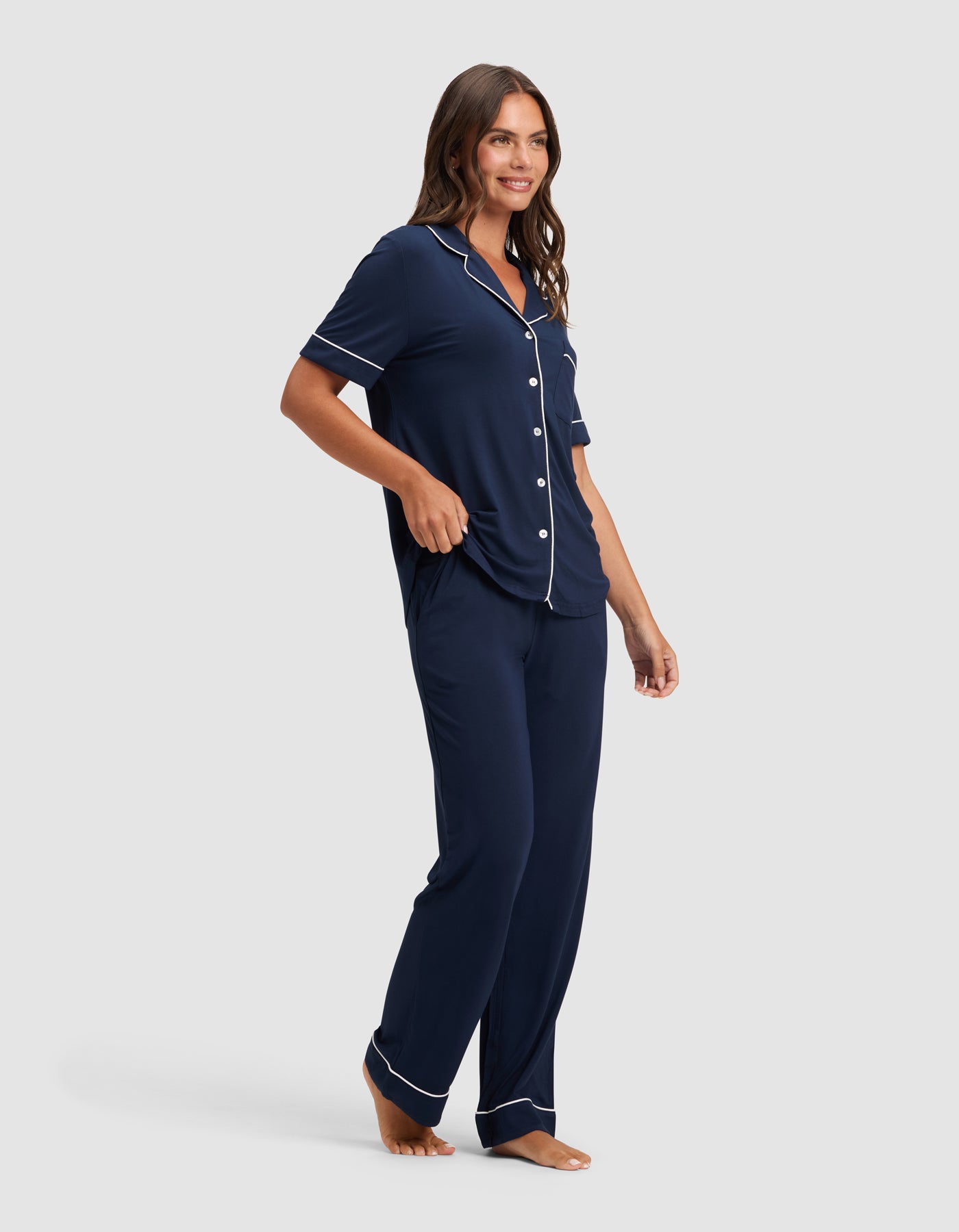 A woman stands barefoot on a light background, smiling and looking aside while wearing Cozy Earth's Women's Bamboo Stretch-Knit Short Sleeve & Pant Pajama Set in navy blue with white piping and a button-up top.