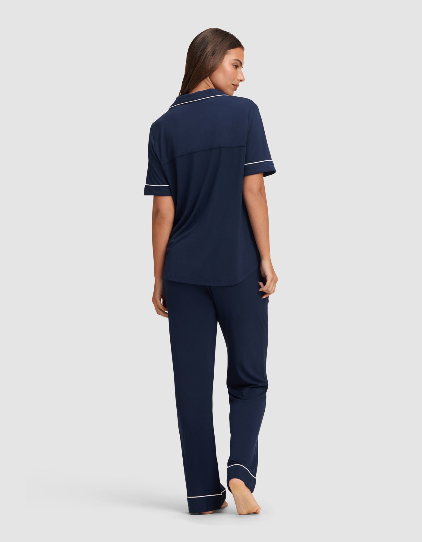 A woman stands barefoot, facing away, wearing the Cozy Earth Women's Bamboo Stretch-Knit Short Sleeve & Pant Pajama Set in navy blue with white trim. The relaxed-fit pajamas have short sleeves and are shown against a plain light gray background.