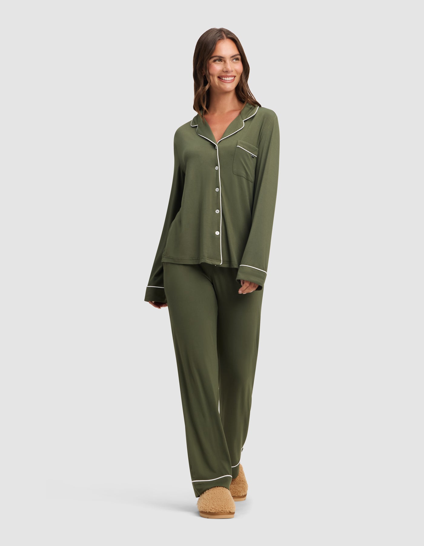 A woman smiles against a light gray background, wearing Cozy Earth's Women's Bamboo Stretch-Knit Long Sleeve Pajama Set in olive green with white piping and beige slippers.