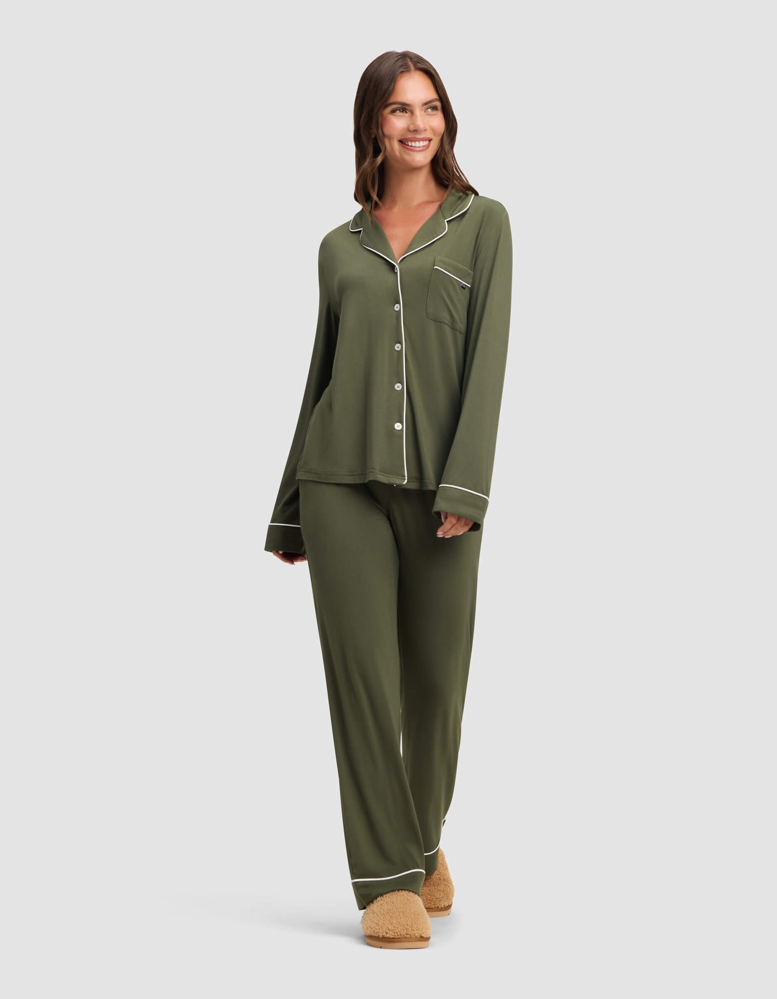 A woman smiles in the Cozy Earth Women's Bamboo Stretch-Knit Long Sleeve Pajama Set in olive green with white piping and tan slippers. The pajama top features buttons and a chest pocket; the loose pants complete her look against a light background.