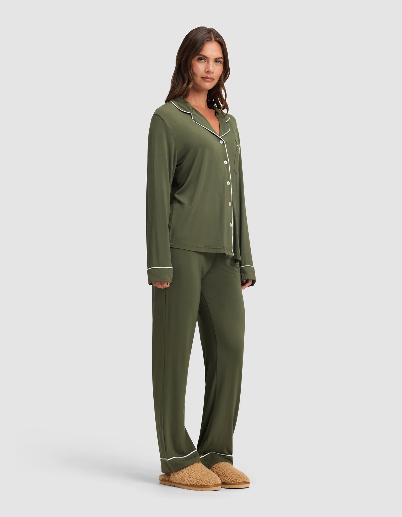 A woman wears the Cozy Earth Women's Bamboo Stretch-Knit Long Sleeve Pajama Set in olive green with matching pants and tan slippers, standing against a plain light gray background. 