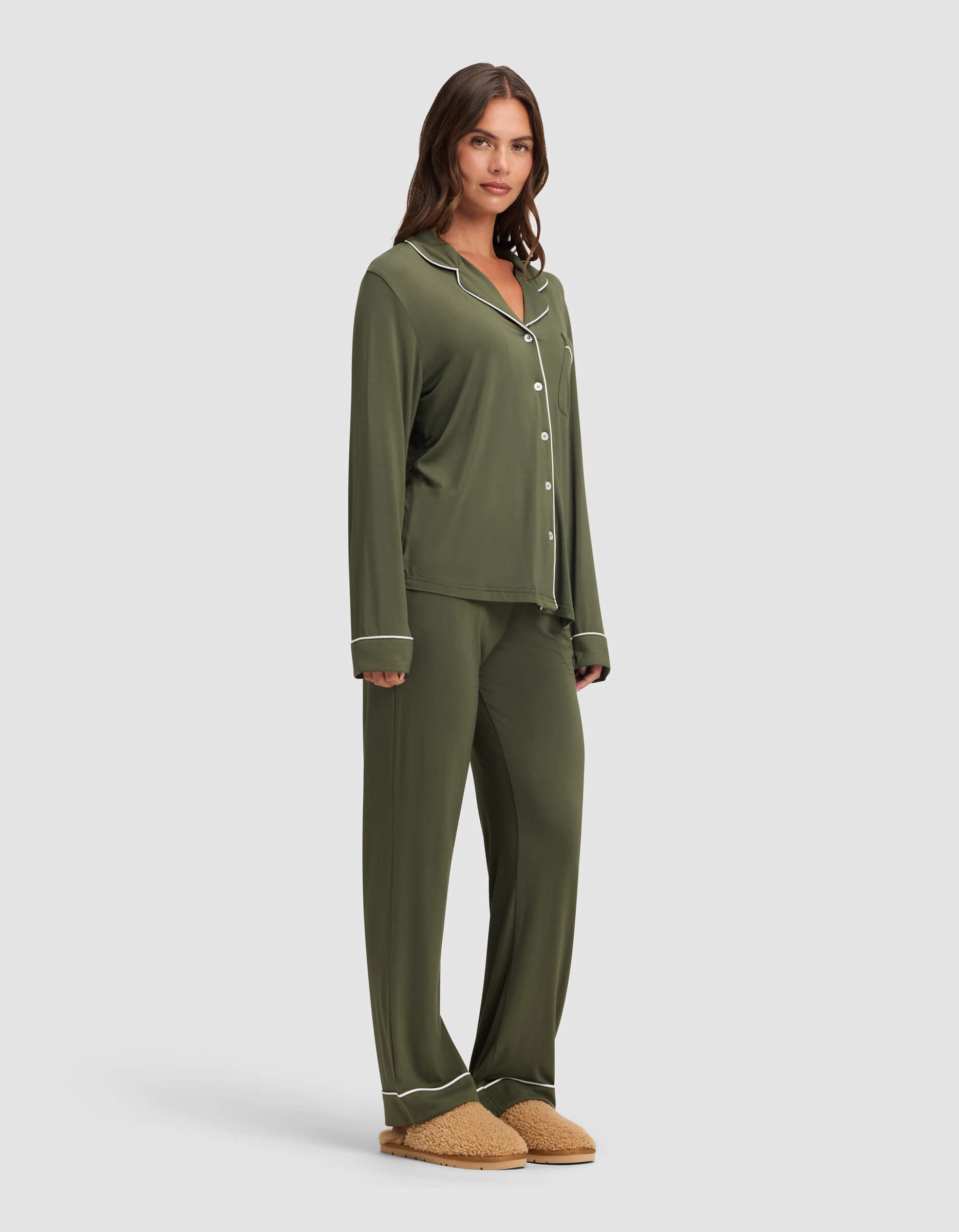 A woman wears the Cozy Earth Women's Bamboo Stretch-Knit Long Sleeve Pajama Set in olive green with white piping, featuring a collared button-up top and loose-fit pants, paired with tan slippers. She has long brown hair and a neutral expression.