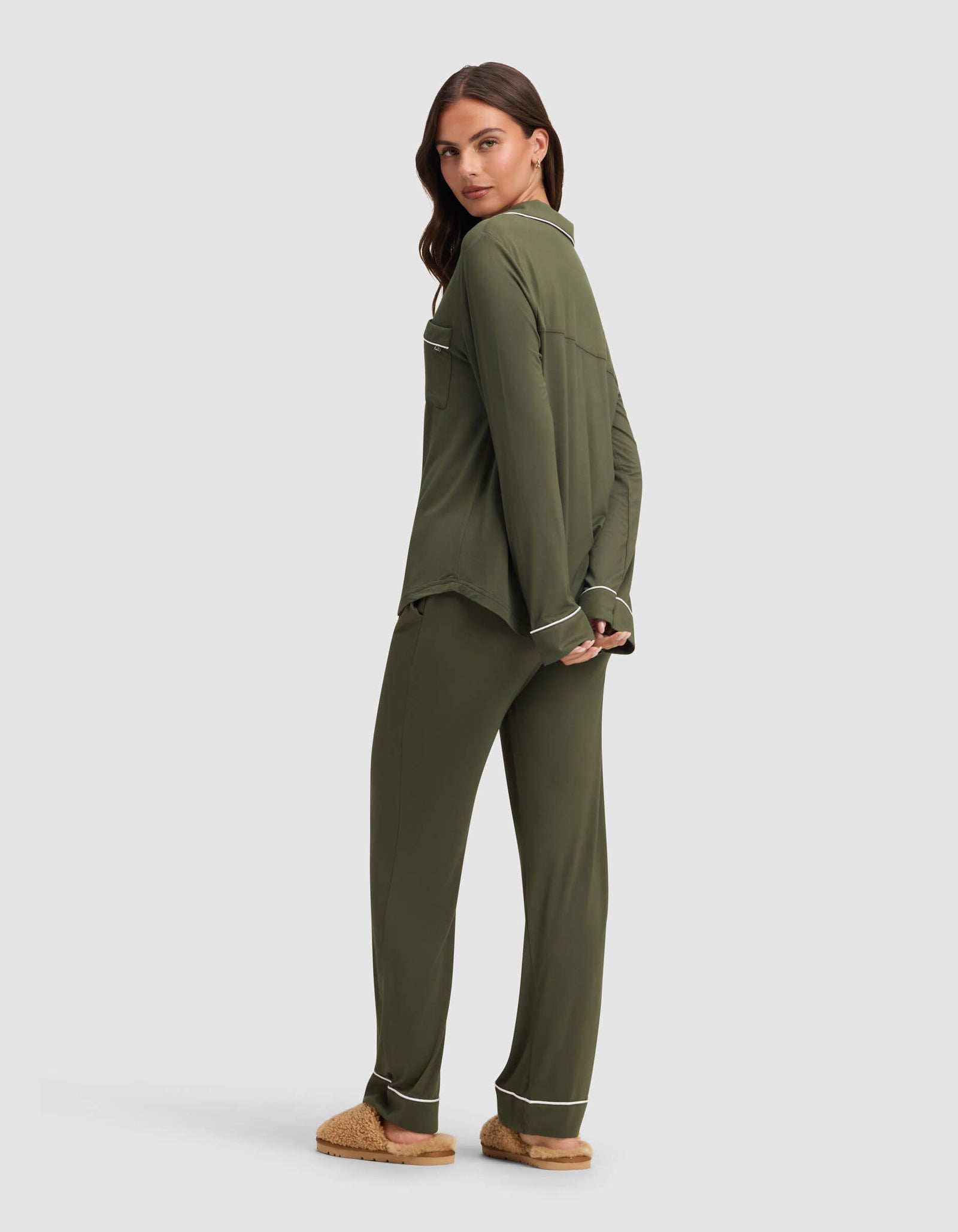 A woman, turned slightly away with her hands behind her back, models Cozy Earth's Women's Bamboo Stretch-Knit Long Sleeve Pajama Set in olive green with white trim and tan slippers against a light gray background.