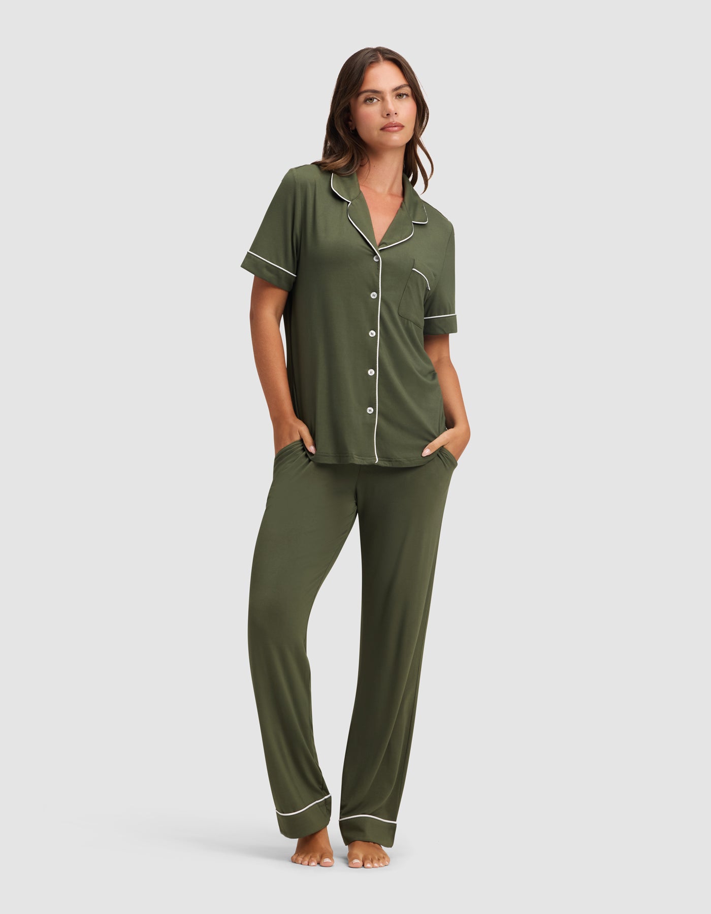 A woman stands barefoot in the Cozy Earth Women's Bamboo Stretch-Knit Short Sleeve & Pant Pajama Set in olive green with white piping, featuring a button-up top and long pants, her hands in her pockets against a plain light background.
