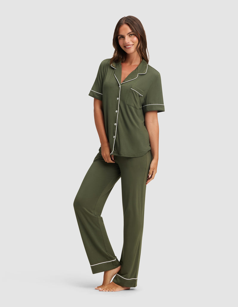 A barefoot woman smiles, wearing Cozy Earth's Women's Bamboo Stretch-Knit Short Sleeve & Pant Pajama Set in olive green with white piping, featuring a button-up top with a chest pocket and matching long pants.