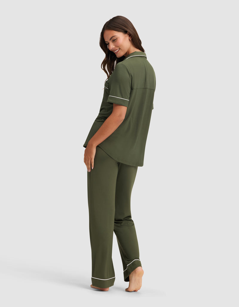 A woman stands barefoot, smiling over her shoulder, wearing the Cozy Earth Women's Bamboo Stretch-Knit Short Sleeve & Pant Pajama Set in olive green with white piping. The background is plain light gray.
