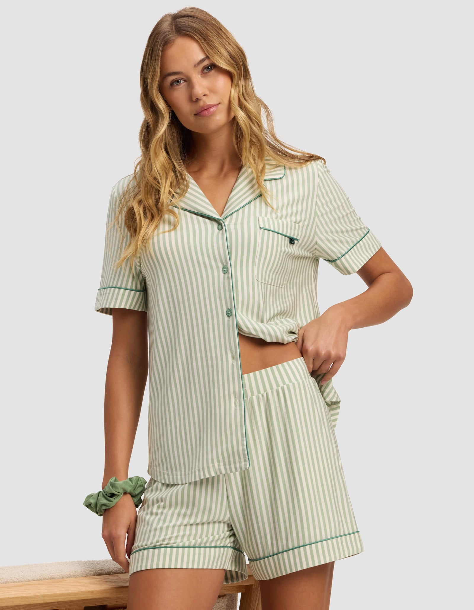 A woman stands indoors wearing Cozy Earth’s Women's Bamboo Stretch-Knit Pajama Short set in light green and white stripes, featuring a short-sleeve top and matching shorts, with one hand on her hip and the other relaxed by her side.