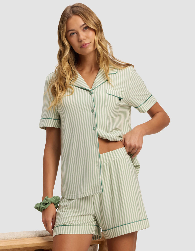 A woman stands indoors wearing Cozy Earth’s Women's Bamboo Stretch-Knit Pajama Short set in light green and white stripes, featuring a short-sleeve top and matching shorts, with one hand on her hip and the other relaxed by her side.