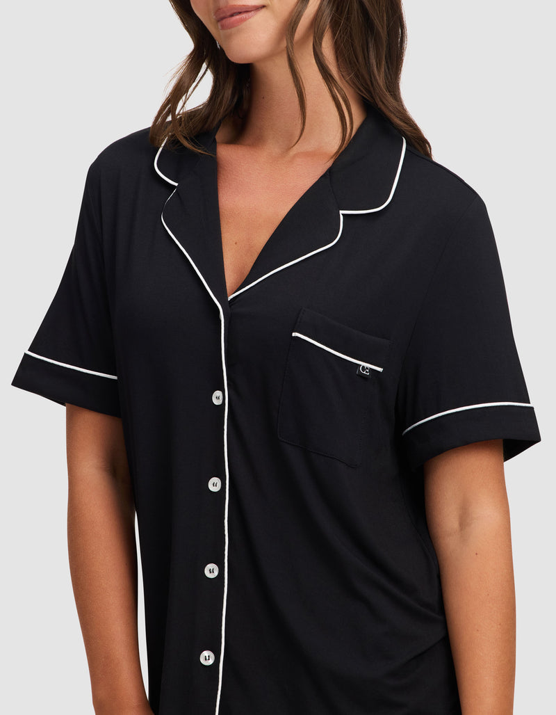 A woman wears the Cozy Earth Women's Bamboo Stretch-Knit Short Sleeve Pajama Set in black, featuring white piping, front buttons, and a chest pocket, standing against a plain light background.