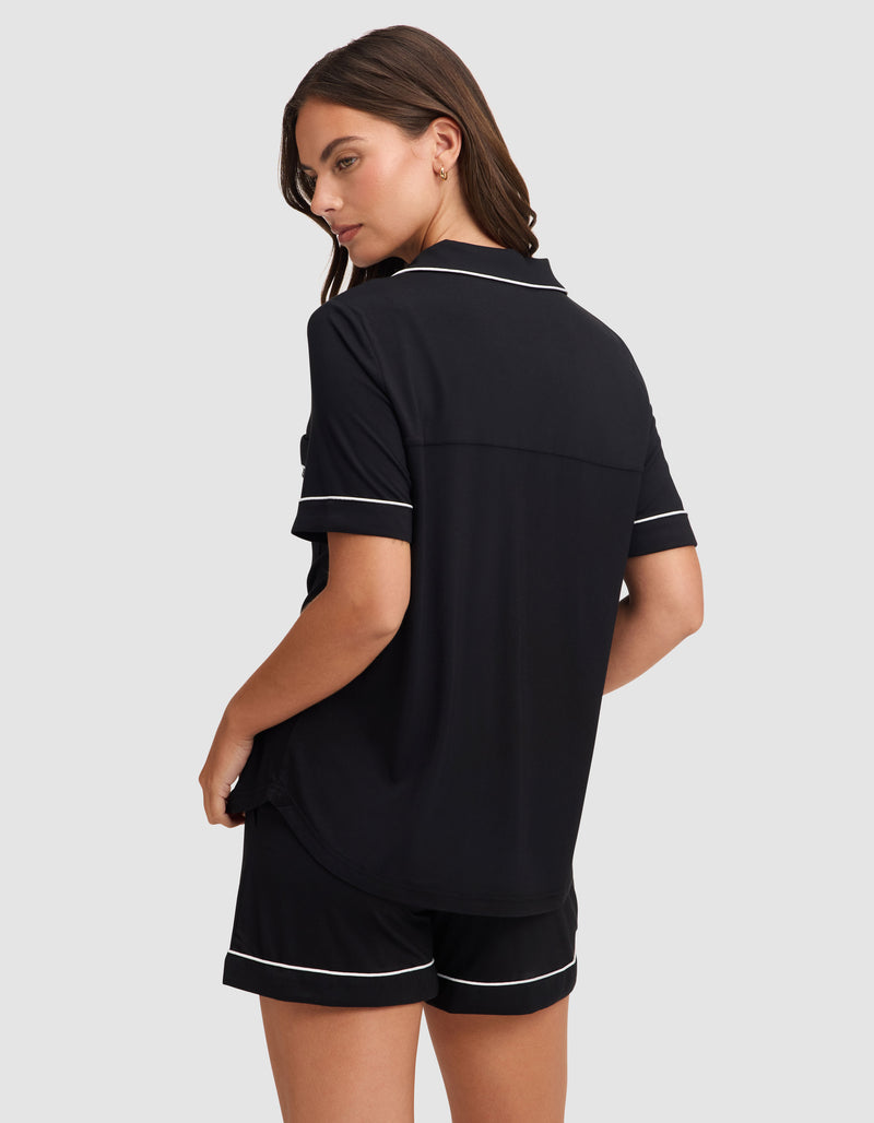 A woman models the Cozy Earth Women's Bamboo Stretch-Knit Short Sleeve Pajama Set in black with white trim, featuring a collared top and matching shorts, while standing against a plain light gray background.