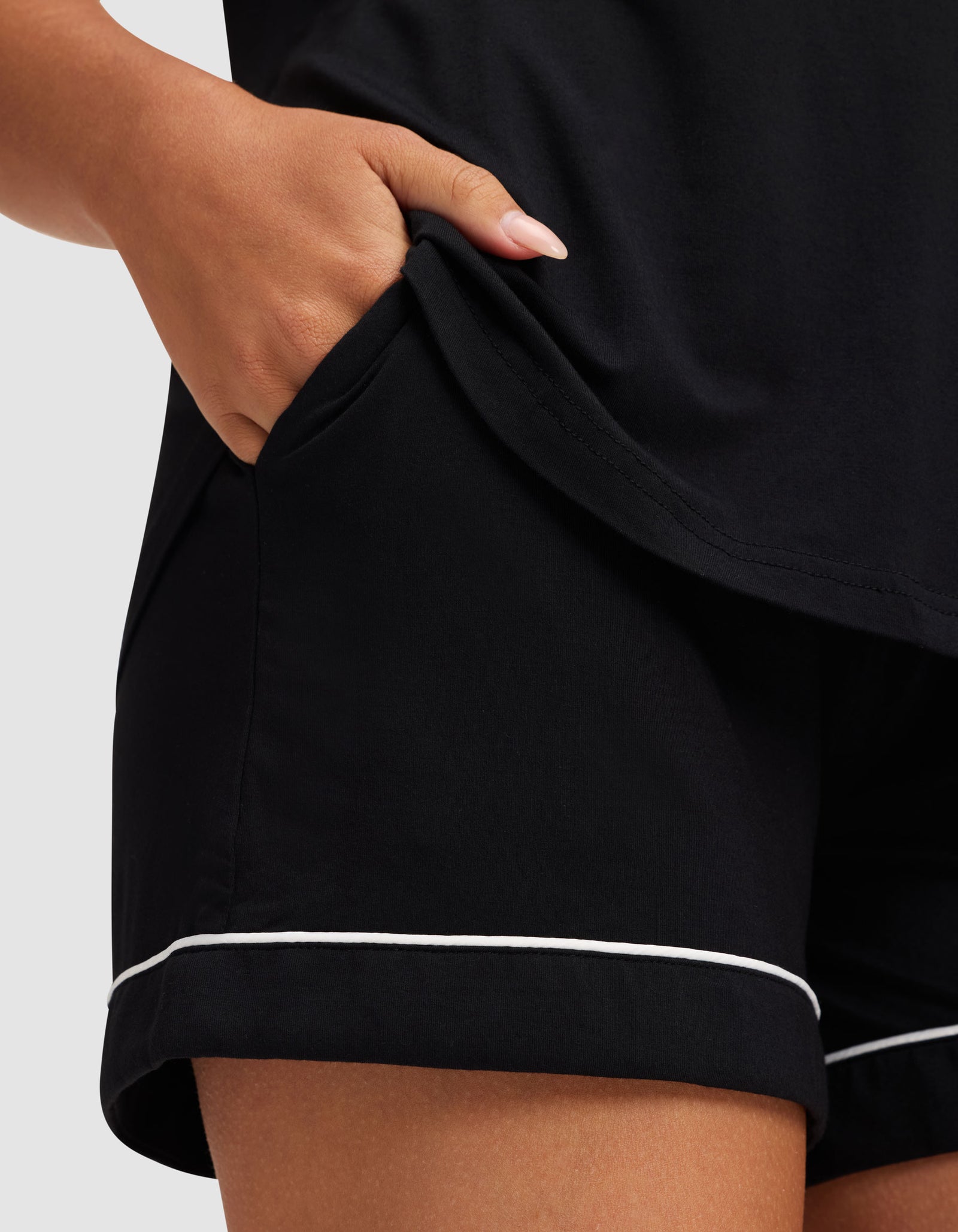 A person’s hand rests in the pocket of Cozy Earth’s Women’s Bamboo Stretch-Knit Short Sleeve Pajama Set—black shorts with white piping and a matching top. Only the lower torso, hand, and part of the outfit are visible.  