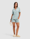 A woman wears the Cozy Earth Women's Bamboo Stretch-Knit Short Sleeve Pajama Set in light blue, paired with matching shorts and beige slippers. She smiles, holding a white tumbler, with one hand in her pocket and a blue scrunchie on her wrist. |Color:Brook