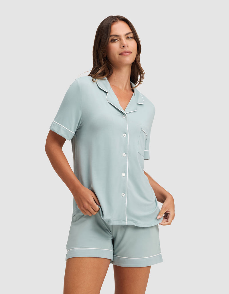 A woman stands against a plain light gray background wearing the Cozy Earth Women's Bamboo Stretch-Knit Short Sleeve Pajama Set in light blue with white trim, featuring a button-up top and matching shorts.