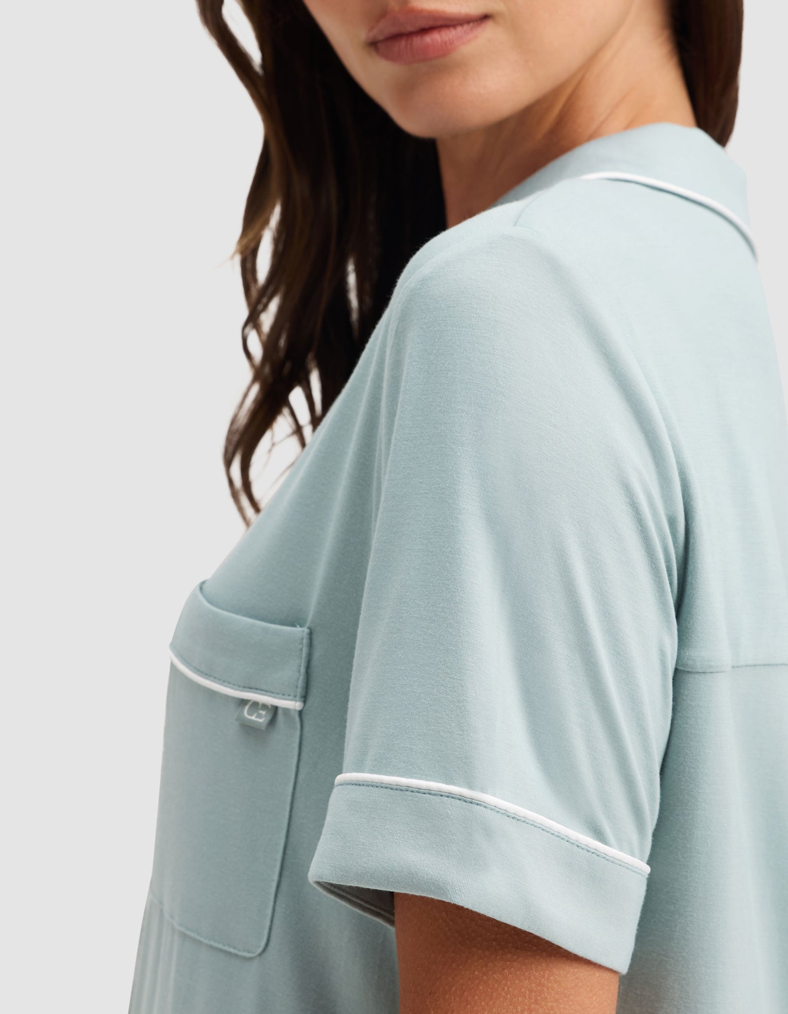 A woman with wavy brown hair models the Cozy Earth Women's Bamboo Stretch-Knit Short Sleeve Pajama Set in light blue, featuring white piping on the pocket and sleeves. The image is cropped from nose to above waist against a plain background. 