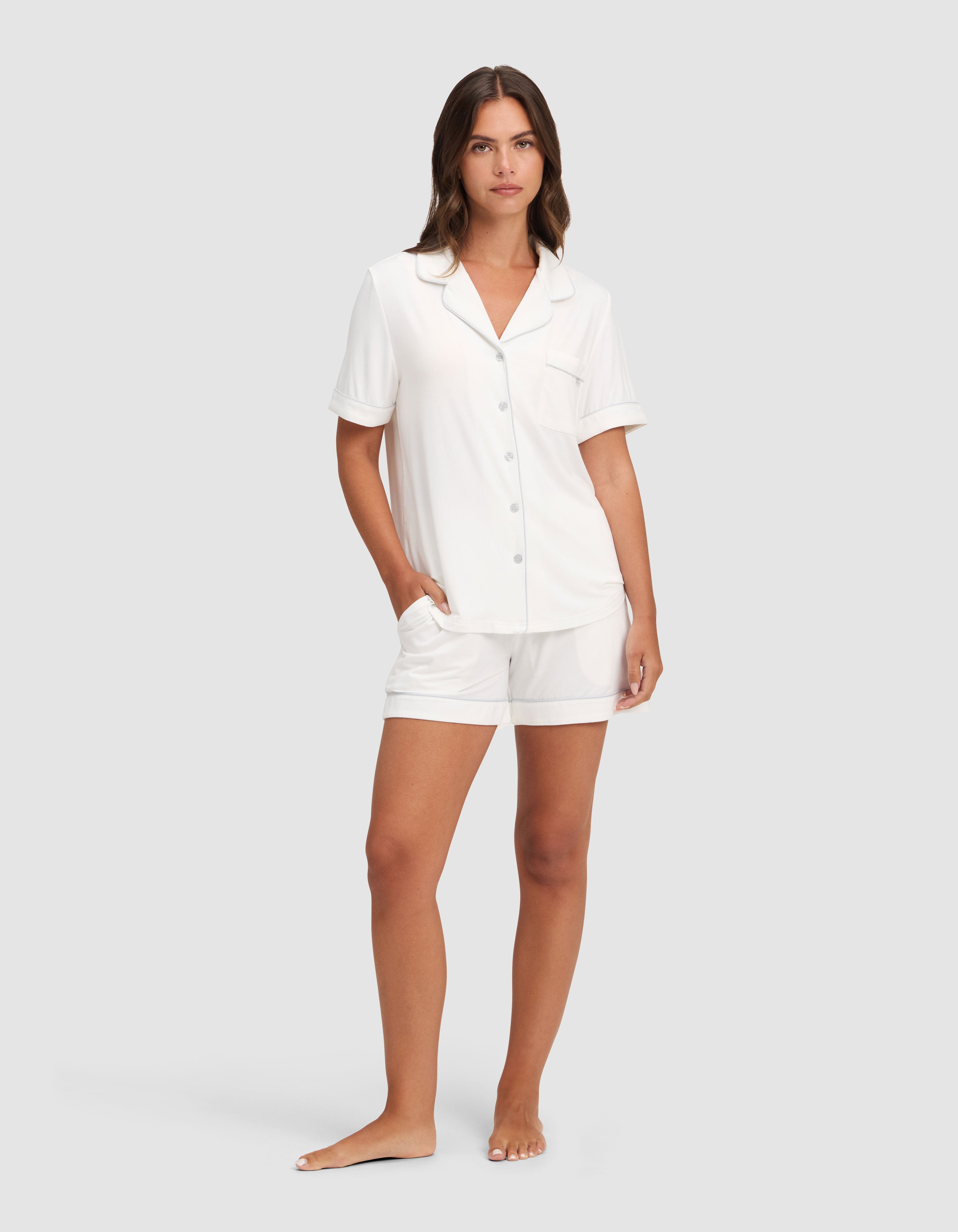 A woman stands barefoot against a plain background, wearing the Cozy Earth Women's Bamboo Stretch-Knit Short Sleeve Pajama Set. One hand is in her pocket as she gazes calmly at the camera. |Color:Ivory